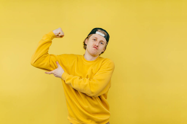 Grappige man in sweatshirt en pet toont biceps op een gele achtergrond en trekt een grappig gezicht. geïsoleerd. - Foto, afbeelding
