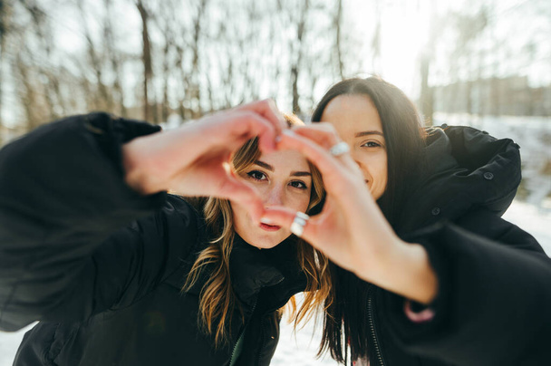 Due ragazze in giacche nere camminano insieme in un parco invernale, guardano la macchina fotografica e mostrano un gesto di cuore, le donne amano le passeggiate invernali. - Foto, immagini