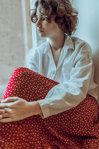 Lonely lovely teen girl is sad. Portrait of bored teenage girl, with brown curly hair, in white men's shirt, colorful red bright skirt, sitting on windowsill by window, clasping legs with hands. Loneliness concept - Photo, Image