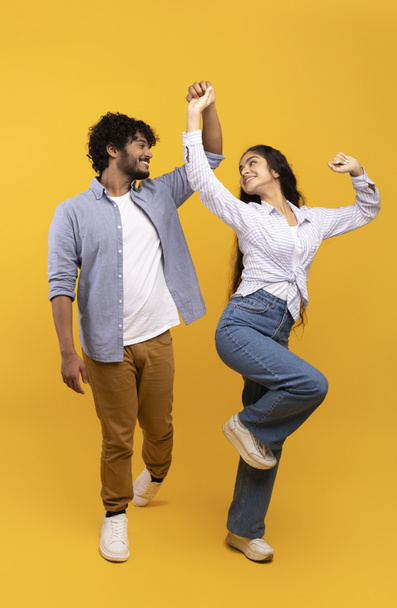 Affectionate indian couple holding hands and dancing together over yellow background, enjoying favourite song, crop - Fotografie, Obrázek