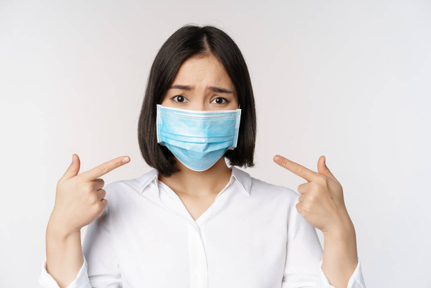 Close up of sad asian girl in medical face mask pointing at her head and looking upset, standing over white background - Photo, image