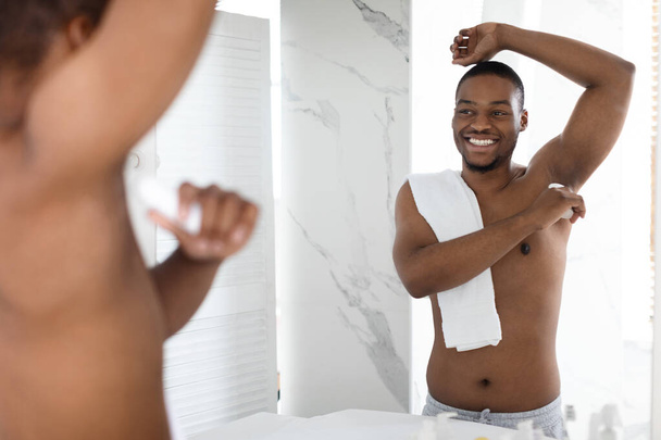 Mladý afroameričan bez trička aplikující deodorant podpaží v koupelně - Fotografie, Obrázek
