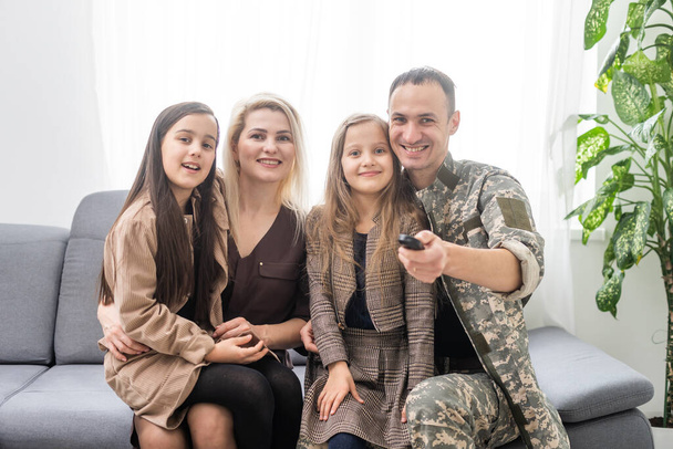 Jonge dappere soldaat die kwaliteitstijd doorbrengt met zijn familie. Ze kijken televisie. Man met afstandsbediening in handen. - Foto, afbeelding