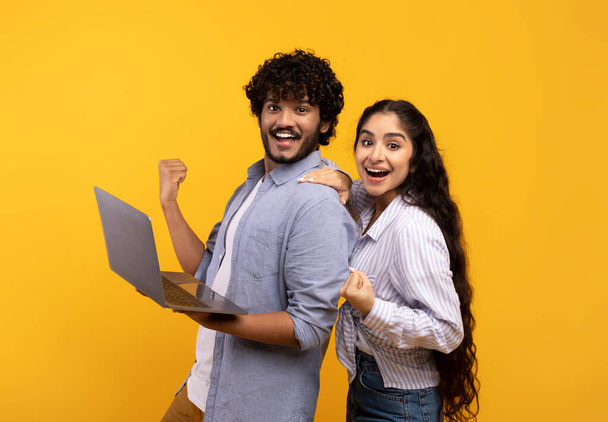 Ah oui ? Couple indien excité tenant un ordinateur portable, acclamant et regardant la caméra, célébrant la victoire, fond jaune - Photo, image