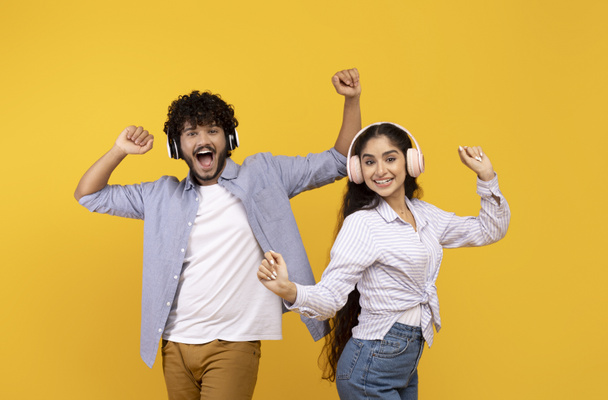 Diviértanse y disfruten la música juntos. Feliz pareja india joven bailando en auriculares, fondo amarillo - Foto, Imagen