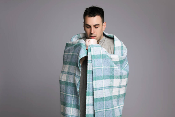 Young man wrapped in blanket with cup of hot drink suffering from cold on grey background - Fotó, kép