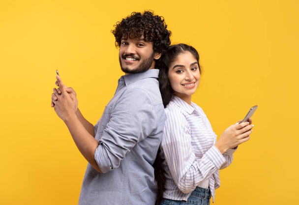 Estilo de vida moderno. Emocionada pareja india de pie espalda con espalda, sosteniendo y utilizando sus teléfonos celulares, sonriendo a la cámara - Foto, Imagen