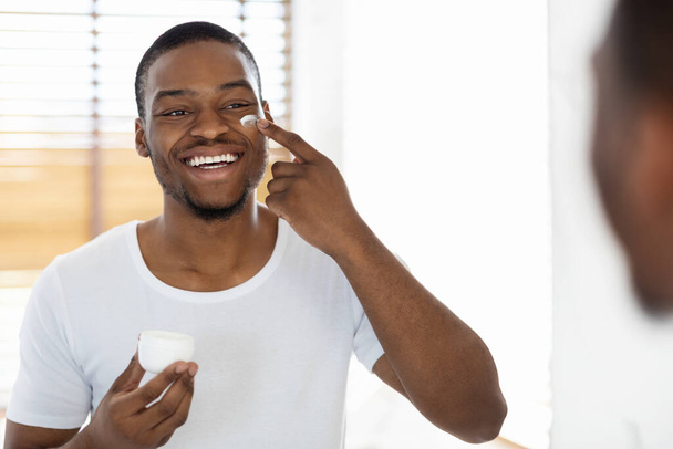 Щасливий чорний чоловік застосовує крем для обличчя біля дзеркала у ванній
 - Фото, зображення