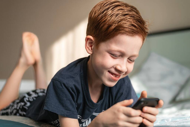 Heureux enfant roux et la technologie numérique, le garçon tient un téléphone dans ses mains écrit des messages ou joue à des jeux vidéo et sourit. - Photo, image