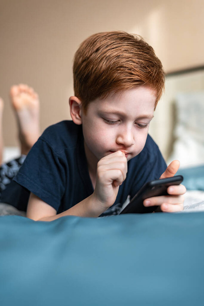Concepto de comunicación y tecnología - chico pelirrojo pensando con smartphone, jugando en casa o enviando mensajes de texto. - Foto, imagen