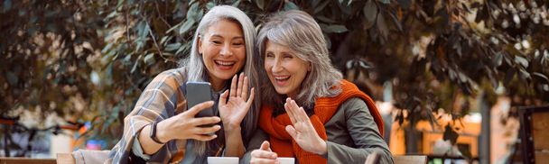 Позитивные зрелые женщины друзья машут руками в видеочате через смартфон в уличном кафе - Фото, изображение