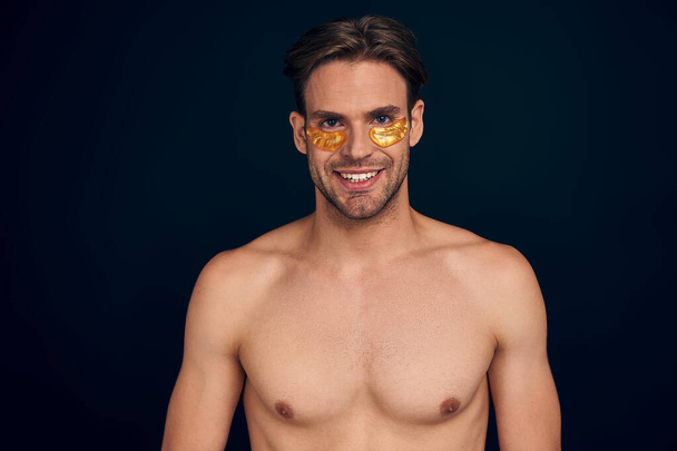 Smiling bearded naked young man 20s perfect skin wearing moisturizing patches under eyes put hand prop up on chin isolated on pink background studio. Skin care healthcare cosmetic procedures concept. - Φωτογραφία, εικόνα