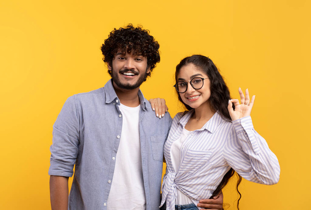 Positive indian spouses embracing and lady showing okay gesture over yellow studio background - Photo, Image