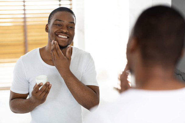 Aftershave Gondozás. Vonzó afro-amerikai srác használ hidratáló krém a fürdőszobában - Fotó, kép