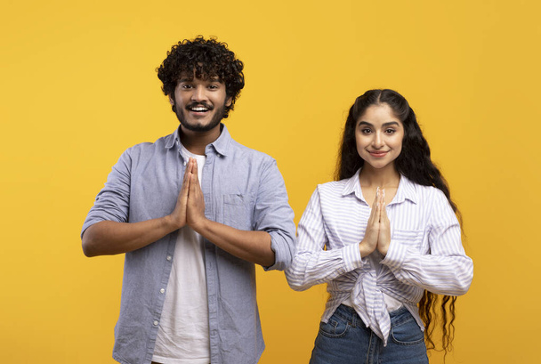 Peaceful indian couple putting hands together in namaste or prayer gesture, looking at camera on yellow background - Photo, Image