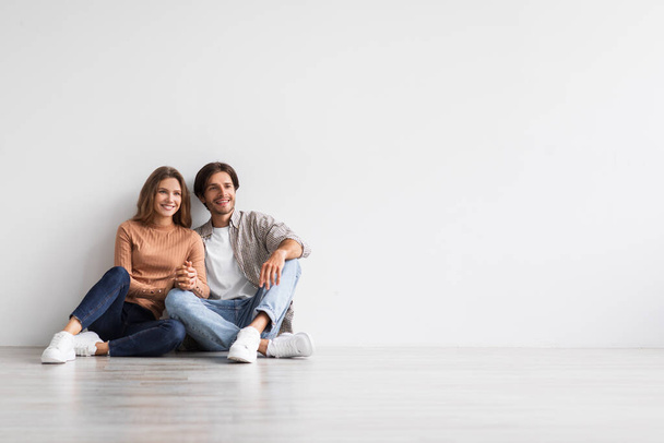 Happy young european couple holds hands, dreaming, planning future interior, sit on floor on white wall background - Foto, Bild