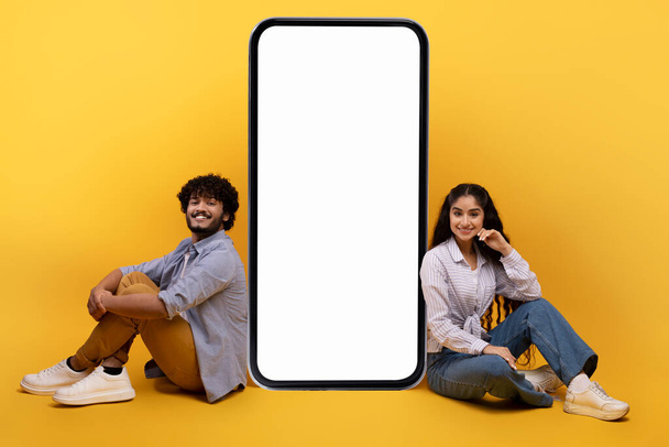 Excited indian couple sitting near giant smartphone with blank white screen, demonstrating copy space for your ad - Foto, immagini
