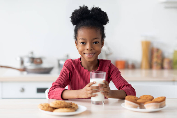 Chica negra alegre sentada en la mesa, disfrutando de la leche y las galletas - Foto, imagen