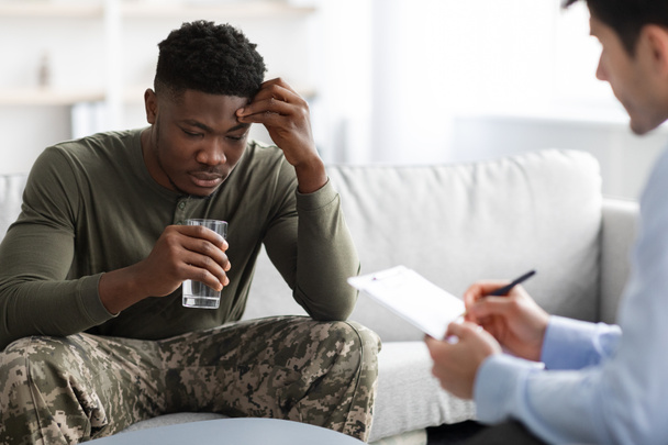 Emotional black military man drinking water while talking to psychotherapist - Photo, Image