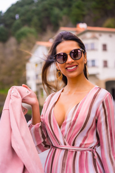 Portrait of a young Latin woman in a pink dress and sunglasses enjoying summer vacations in a beautiful coastal town, posing by the sea - Fotografie, Obrázek