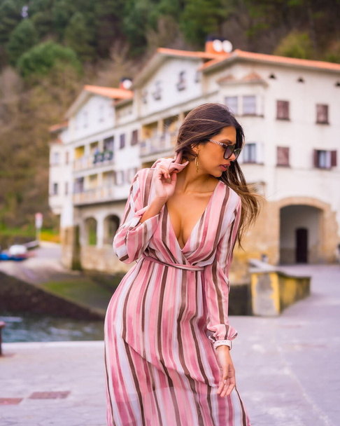 Portrait of a young Latin woman in a pink dress and sunglasses enjoying summer vacations in a beautiful coastal town, posing fashionably by the sea, vertical photo - Valokuva, kuva
