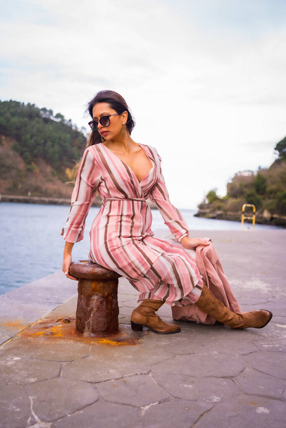 Portrait of a young Latin woman in a pink dress and sunglasses enjoying summer vacations, sitting by the sea - Fotografie, Obrázek