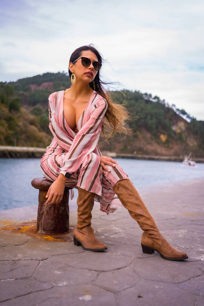 Portrait of a young Latin woman in a pink dress, brown boots and sunglasses enjoying summer vacations, sitting by the sea, vertical photo - Foto, Imagem