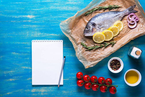 Риба Дорадо на кулінарному папері зі спеціями та блокнотом для рецептів або меню. Вид зверху на синьому фоні
. - Фото, зображення