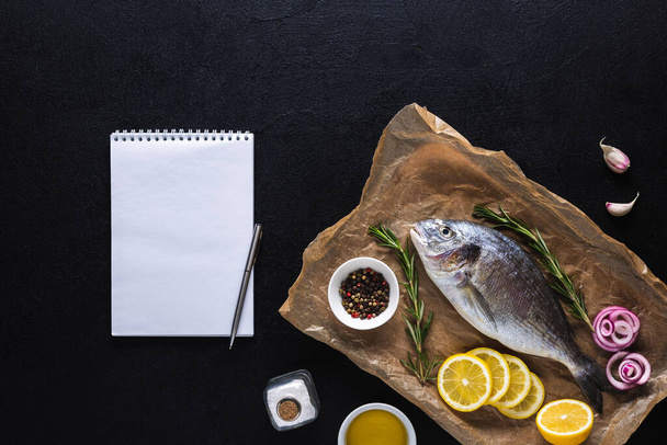 Nyers Dorado főzés papír fűszerek és egy notebook recept vagy menü. Egészséges étel, tenger gyümölcsei. Top nézet egy fekete háttér. - Fotó, kép