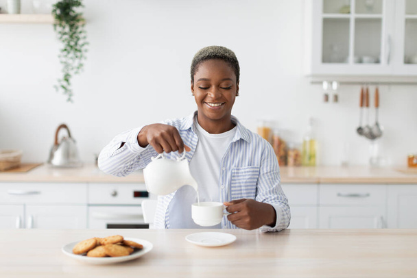 笑顔千年アフリカ系アメリカ人女性でカジュアルな注ぎ牛乳にコーヒーでテーブルミニマリストキッチンインテリア - 写真・画像