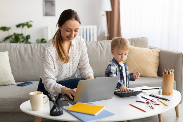 Trabajando como autónomo en la licencia de maternidad. Joven madre trabajando en el ordenador portátil mientras su hijo pequeño jugando con la calculadora neaby - Foto, Imagen