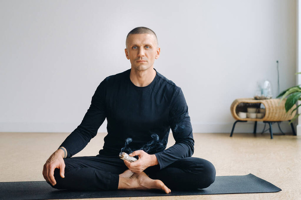 A male athlete in black sportswear sits in the lotus position in his hands with an incense candle in the gym. - Foto, imagen