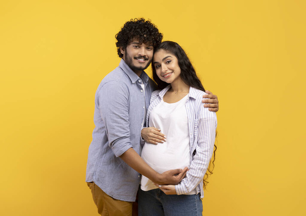 Zufriedener junger indischer Mann umarmt schwangere Frau und berührt Bauch, lächelt gemeinsam in die Kamera vor gelbem Hintergrund - Foto, Bild