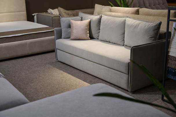 Выставка мягких стильных удобных светло-серых диванов с розовыми подушками возле стенда с образцами ткани и образцами различных текстур и качеств. Мебельный салон - Фото, изображение