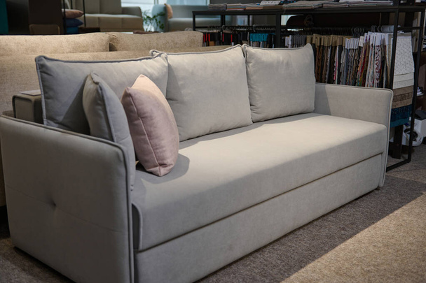 Exposición de elegante sofá acolchado gris claro cómodo con almohadas de color rosa cerca de un soporte con muestras de muestras de telas y muestras de diferentes texturas y calidades. Muebles tienda showroom - Foto, imagen