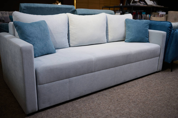 Hermoso sofá minimalista elegante gris claro con cojines azules, que se muestra a la venta en la sala de exposición de la tienda de muebles. Salón amueblado con elegantes muebles tapizados modernos y confortables. - Foto, imagen