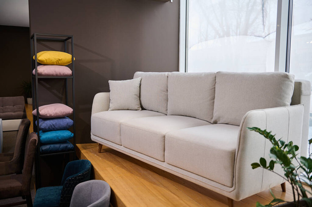 Μοντέρνος μινιμαλιστικός λευκός καναπές με μαξιλάρια προς πώληση σε εκθεσιακό χώρο μοντέρνων επίπλων, δίπλα στο περίπτερο με πολύχρωμα μαξιλάρια από υφάσματα διαφορετικής υφής, απαλότητας, χρωμάτων και ποιότητας - Φωτογραφία, εικόνα