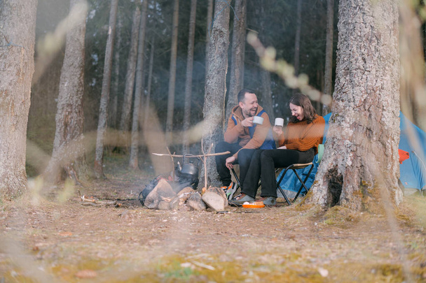 Couple familial se repose sur la rive du lac avec une tente. Fermeture du camp avec un feu dans la forêt. Un homme et une femme secouent le silence par le feu avec des cercles de boissons chaudes - Photo, image