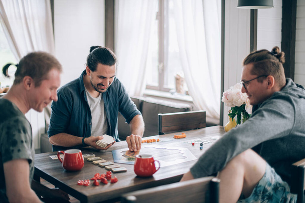 Przyjaciele grają w grę planszową w salonie. Kompania młodych facetów siedzi przy stole i emocjonalnie i radośnie gra w karty w weekend - Zdjęcie, obraz