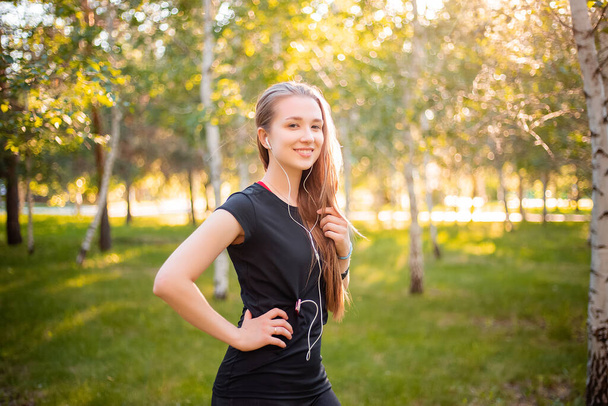 Portrait of a beautiful smiling girl in a sports uniform with flowing hair listening to music in headphones on the background of the park. - Foto, Bild