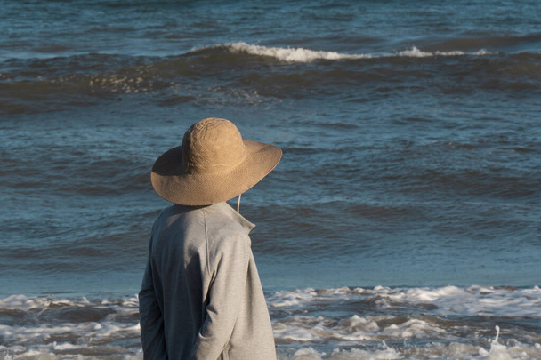 Starsza kobieta, od tyłu patrząca w stronę morza. Ma kapelusz, w tle widać morze z pięknym niebieskim kolorem i falami. - Zdjęcie, obraz