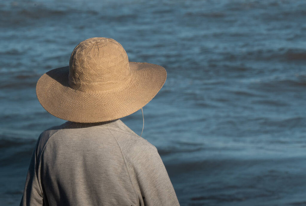 zblízka starší dámu v klobouku s výhledem na moře za slunečného odpoledne. Dáma je na zádech a má na sobě šedou mikinu.. - Fotografie, Obrázek