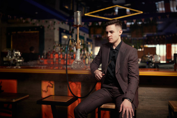 A  handsome well-groomed young man in an expensive suit smoking hookah at the bar in a restaurant. Business person. Luxury lifestyle. - Foto, Imagen