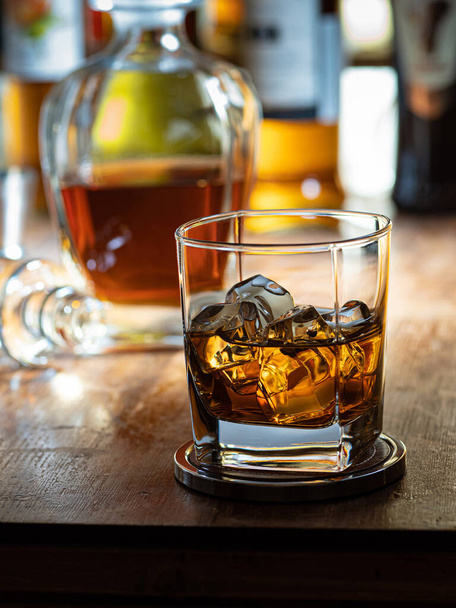 Glass of whisky on the rocks on a wooden bar top with bottles in the background - Foto, afbeelding