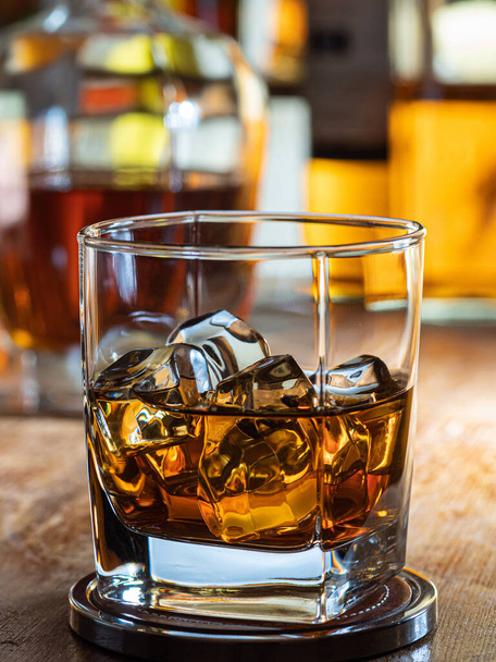 Glas whisky met ijs op een houten barblad met flessen op de achtergrond - Foto, afbeelding