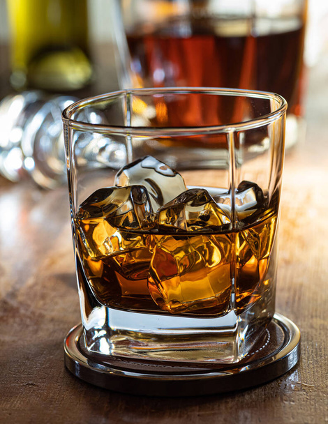 Glass of whisky on the rocks on a wooden bar top with bottles in the background - Foto, imagen
