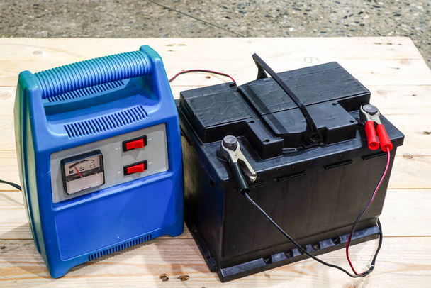 chargeur de batterie bleu câblé à une batterie de voiture noire sur une palette en bois, la charge est en cours - Photo, image