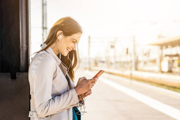 Uśmiechnięta kobieta biznesu korzystająca ze smartfona podczas oczekiwania na dworcu kolejowym - Atrakcyjna młoda studentka korzystająca z telefonu komórkowego stojąca na peronie dworca kolejowego - Zdjęcie, obraz