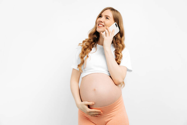 Phone call. Smiling beautiful pregnant woman talking on a mobile phone, consulting a doctor, on an isolated white background with free space - Fotografie, Obrázek