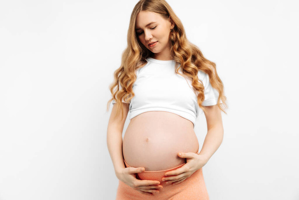 Pregnant happy beautiful woman touches her belly. Pregnant mom caressing her belly and smiling close up. Healthy pregnancy concept, pregnant woman on isolated white background - Φωτογραφία, εικόνα